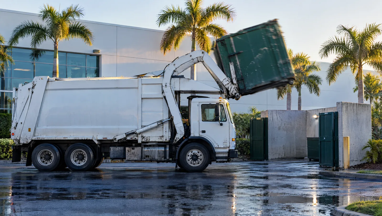 A front-load truck emptying a commercial container behind a Florida office building at sunrise